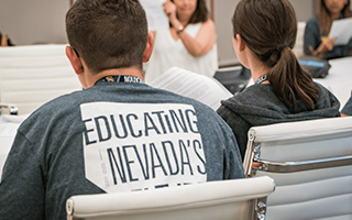 Two students seated for a lecture are pictured from behind. They wear NSC lanyards around their necks.