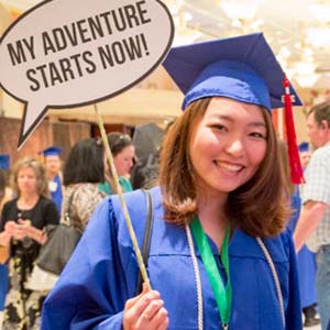 A graduate in a blue cap and gown smiles for the camera. She holds a prop that looks like a speech balloon that contains the words "My Adventure Starts Now."