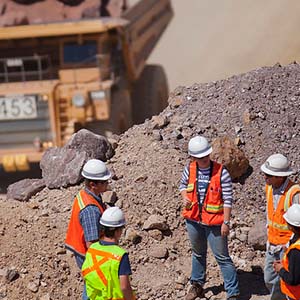 A group of people in reflective vests and hard hats meet in a dirt lot. A construction vehicle can be seen behind them.