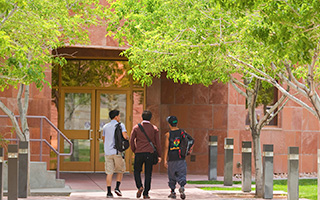 Three students walking shoulder to shoulder are pictured from the rear as they walk towards a red building.