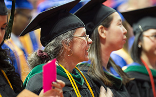 A smiling gray haired woman in a graduate's cap and gown sits amongst an audience of other graduates.