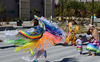 An indigenous American dancer in brightly colored ceremonial dress twirls in the center of a plaza on the CSN campus.