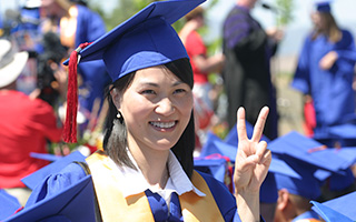 A close-up of a woman in a blue graduation gown who smiles while giving a peace sign to the camera.