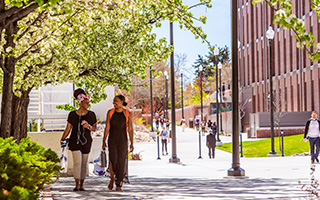 Two students are seen in the foreground walking through the UNR quad on a bright sunny day. They smile warmly at one another. Many other students are seen in the background.