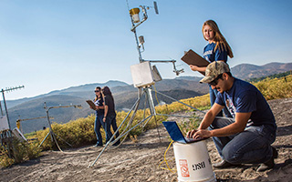 A group of UNR students work with antennae and other technical equipment atop a hill. Rolling tree-covered hills can be seen behind them in the distance.