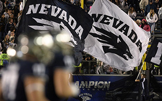 Two flags, one blue and one white and emblazoned with the UNR logo, high across a stadium crowd.