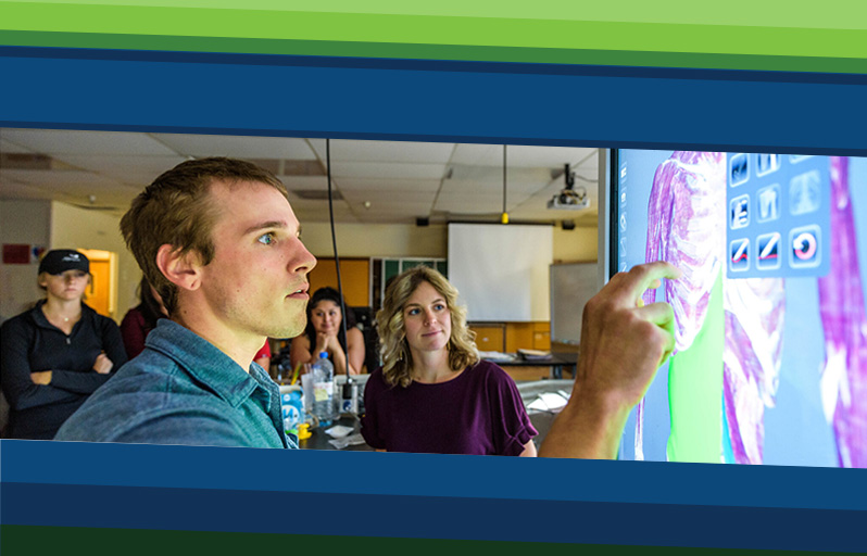 A young man in a classroom works with a touchscreen that displays anatomical data.