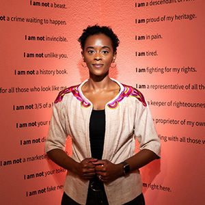 A black woman stands in front of an exhibit with black text on a well-lit red wall.