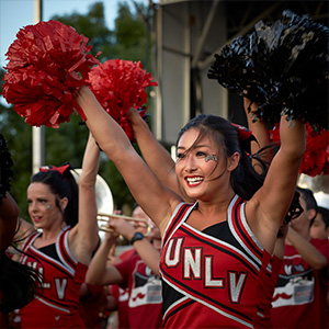 UNLV Cheerleaders hold red and black pop-poms high in the air at an outdoor event.