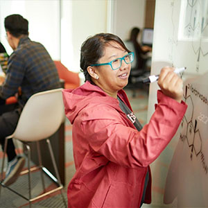 A student in a red jacket adds to scientific notes on a whiteboard.