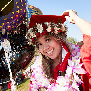A graduate wearing a lei and a ring of flowers around her grad cap smiles while holding a mylar balloon.