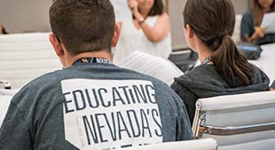 A classroom where two students in the foreground listen intently to a presentation led by a woman a few feet away. The words "Educating Nevada" can be seen clearly on the back of one student's shirt.