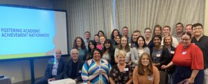 FAAN Convening A large group of people pose for the camera in a meeting room. A screen nearby displays a slide labeled Fostering Academic Achievement Nationwide.