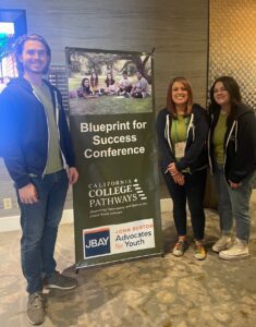 Adam, Laura, & Fia_SIGN2 Three people pose next to a banner stand. The banner reads Blueprint for Success Conference
