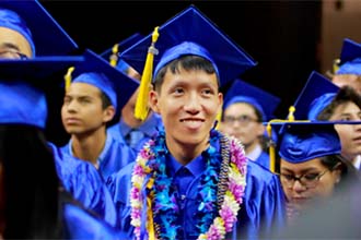 A smiling CSN graduate wearing a lei over his blue cap and gown amidst several other graduates