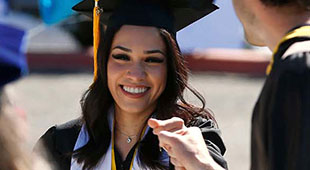 A smiling graduate in a black cap and gown gives a fist bump to a person outside of the frame