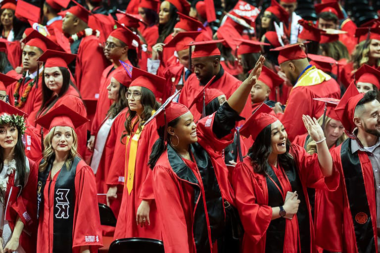 A diverse group of graduates stand together in red graduation robes. Two woman in the front of the crowd wave excitedly to someone off-camera