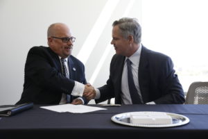 Men in business suits shake hands while seated next to each other at a table draped in blue
