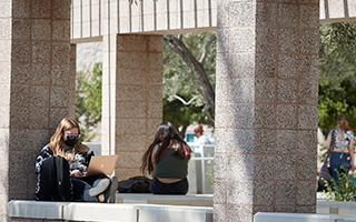 Two students sit outside the UNLV library. The student in the foreground works on a laptop while wearing a facemask.