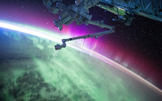 The earth seen from space, a purple aurora is seen around the arc of the earth's curvature. A Robotic arm from a spacecraft can bee seen in silhouette in the foreground.