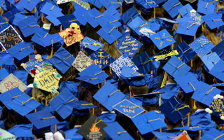 A crowd of graduates in blue caps seen from above. Many of mortarboards are decorated and personalized.
