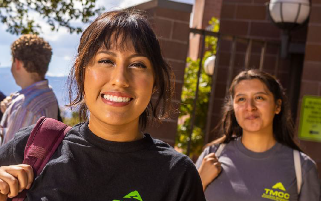 A young woman smiles and faces the viewer. Behind her is another young woman and a group of young men engaged in lively conversation.